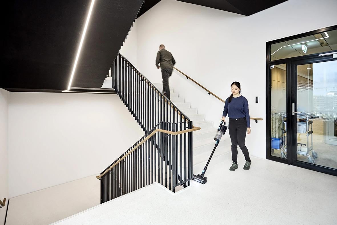 " personer i en modern inomhusmiljö, en går uppför trappan och en annan städar golvet med en dammsugare"