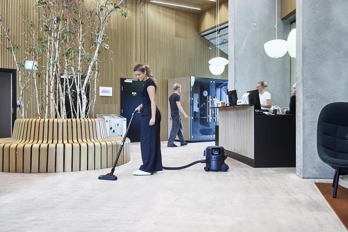 Person som städar med dammsugare i ett modernt inomhusmiljö, soffor och reception i bakgrunden