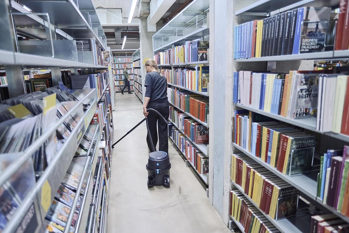 Person städar bibliotek med dammsugare mellan bokhyllor