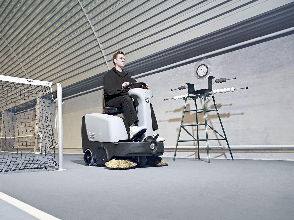 Person kör en Nilfisk SR1000S sopmaskin inomhus på tennisplan