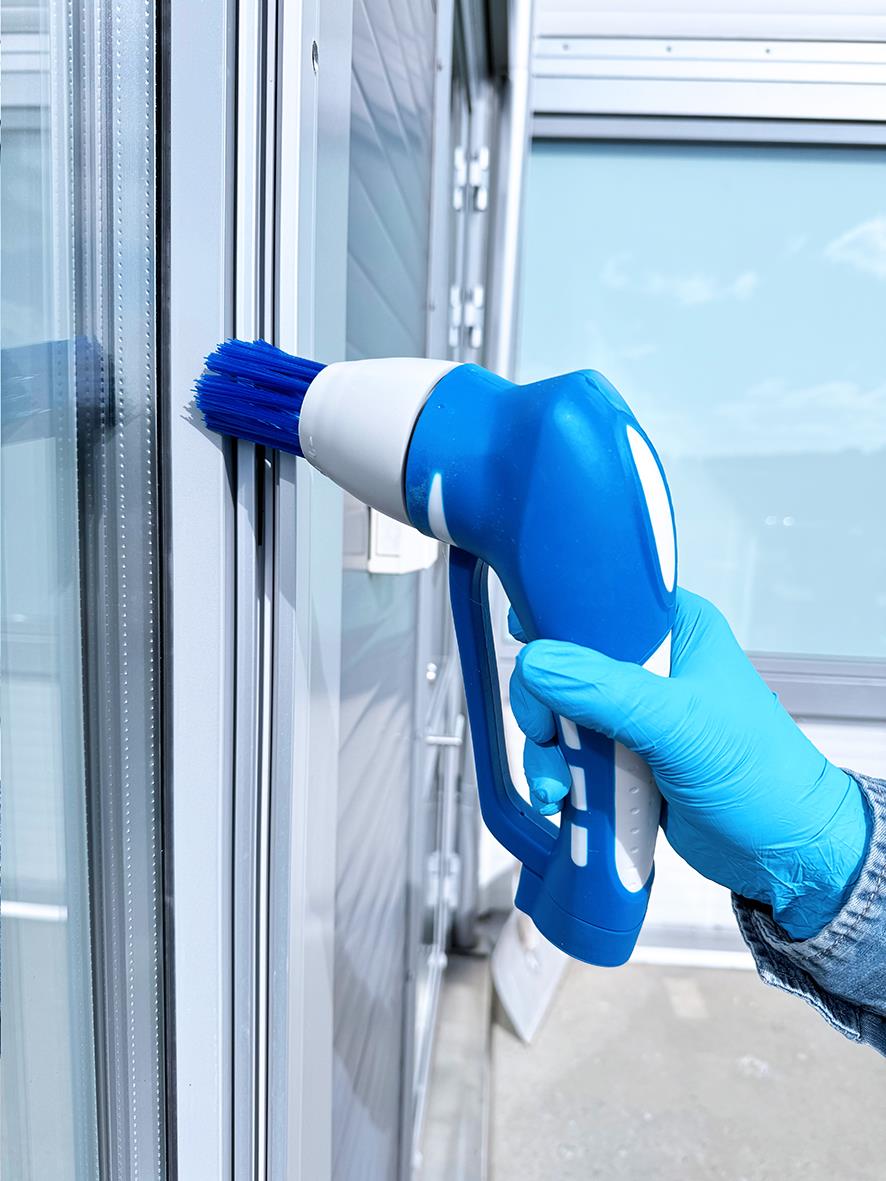 Person i blå handskar använder en blå och vit handhållen skurmaskin för att rengöra en vägg eller yta.