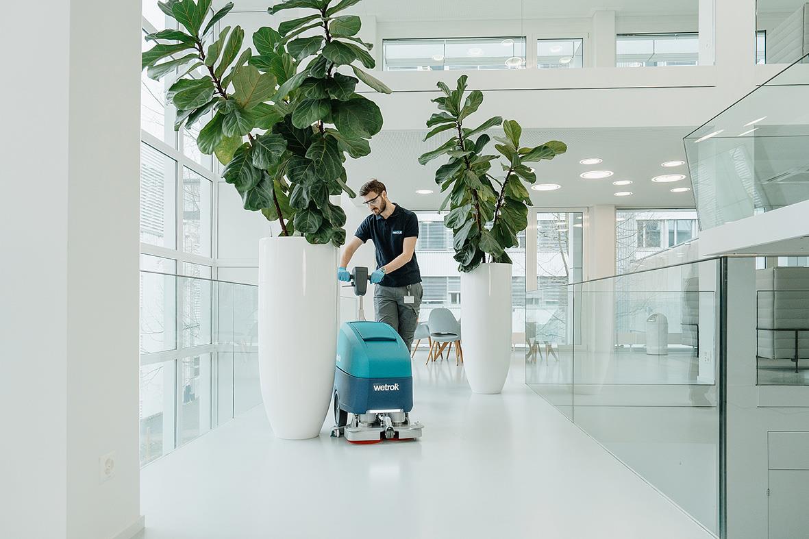 Person som använder kombiskurmaskin Wetrok Mambo med autodosering i ett modernt, ljust kontor med stora växter