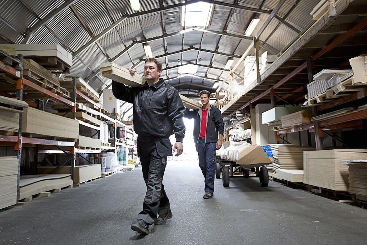 Två män i arbetskläder går genom en lagerlokal med hyllor och byggmaterial