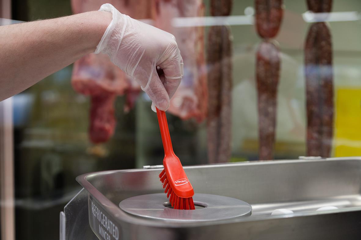 Person håller i en diskborste med röd skaft och borsthuvud, rengör en diskbänk i ett kök eller tvättrum