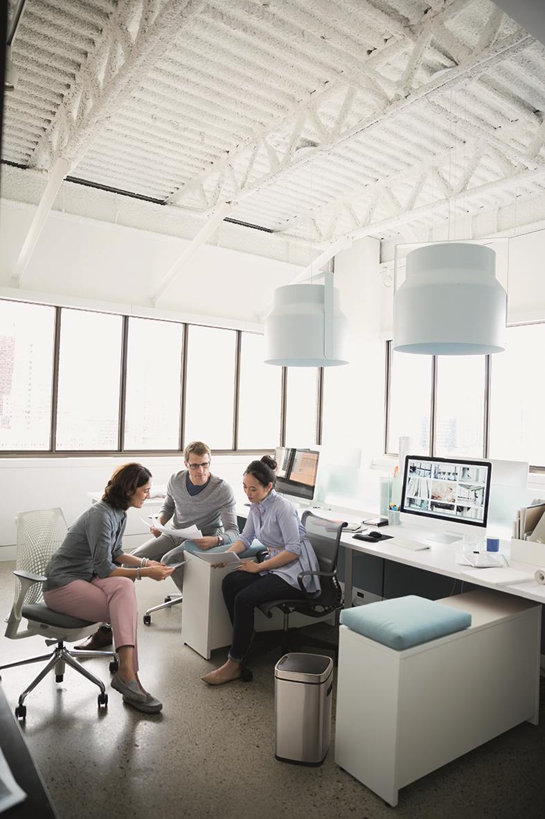 Tre personer i ett kontorsmöte i ett ljust rum med stora fönster, skrivbord med datorer och en papperskorg