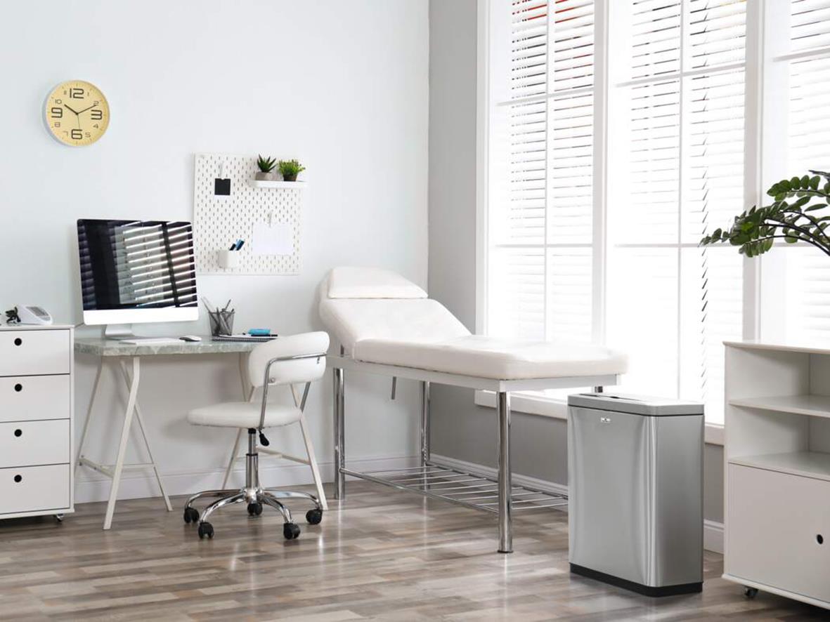 Vårdrum med skrivbord, stol, papperskorg och underskåp i ett ljust rum med stora fönster och växter