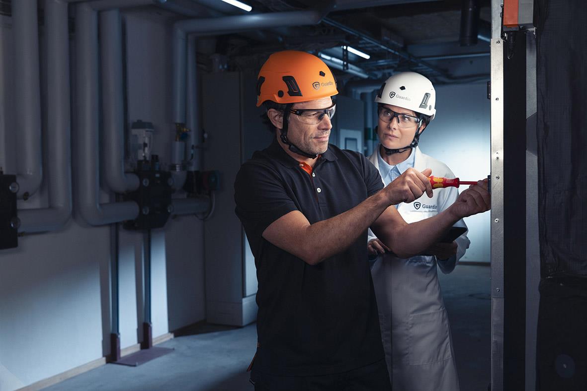 Två personer i skyddsglasögon och hjälmar arbetar med elektrisk utrustning i industrimiljö