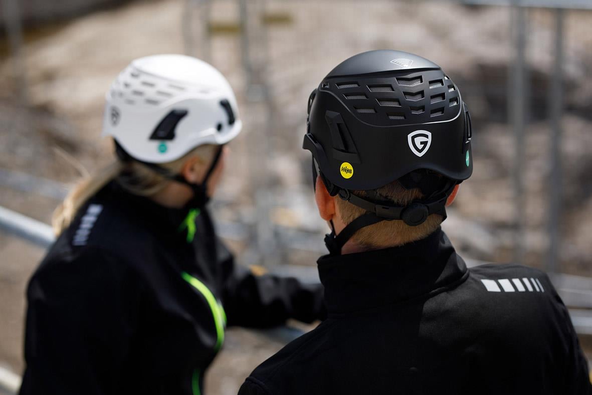 Två personer i hjälmar, en svart Guardio Armet hjälm och en vit hjälm, utomhus i en byggarbetsmiljö