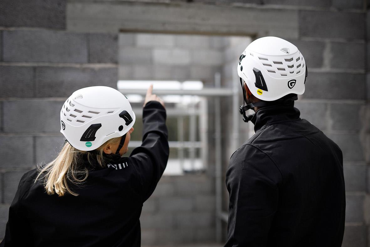 Två personer i skyddshjälmar pekar mot en byggnad, en i vit hjälm och den andra i svart klädsel