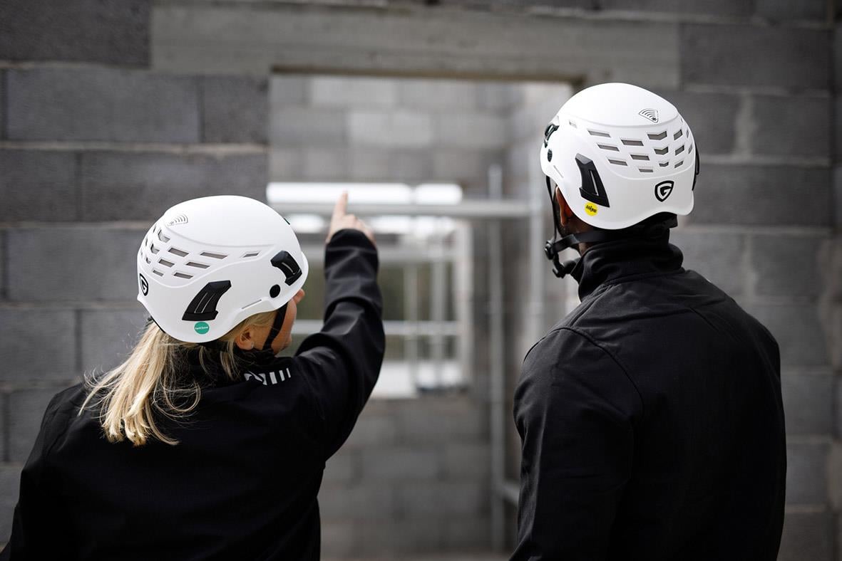 Två personer i skyddshjälmar pekar mot en byggnad, en i vit hjälm och den andra i svart klädsel