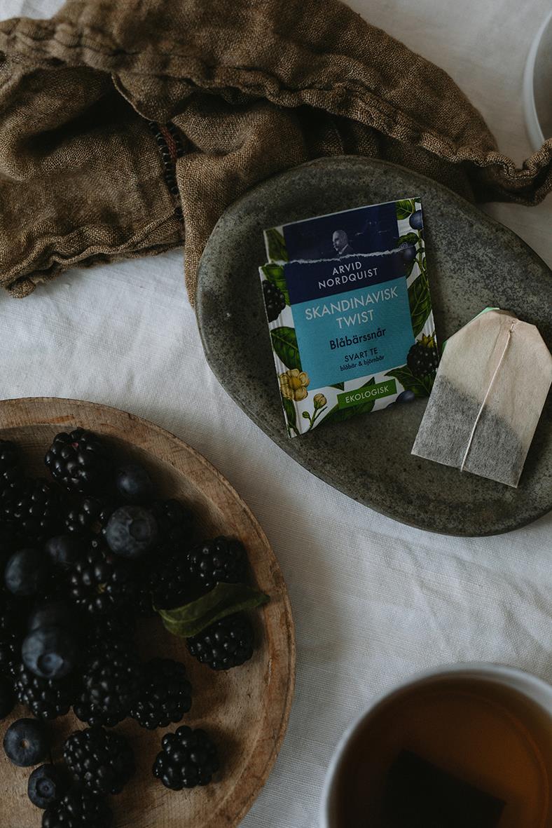 Svart te från Arvid Nordquist med blåbärssnår, omgiven av färska björnbär och blåbär på träfat, med en kopp te i hörnet