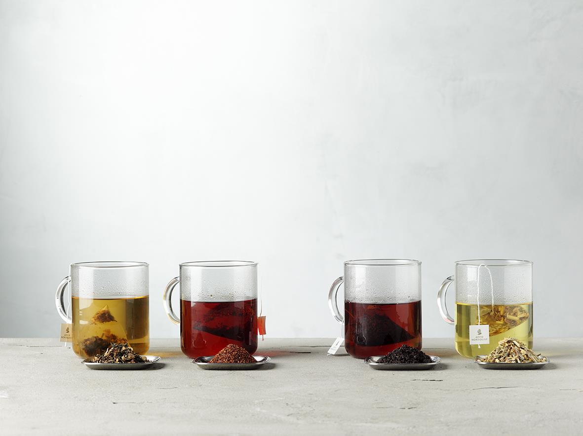 Fyra glas med svart te och olika bär, inklusive blåbärssnår, på en ljus bakgrund