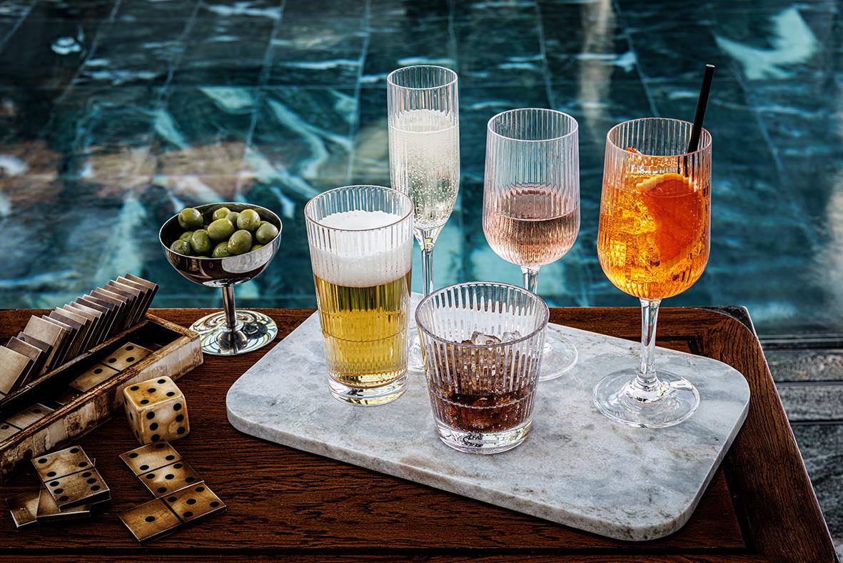 Flera drinkglas av Tritan med olika drycker på en marmorskiva, omgiven av spelkort och gröna oliver