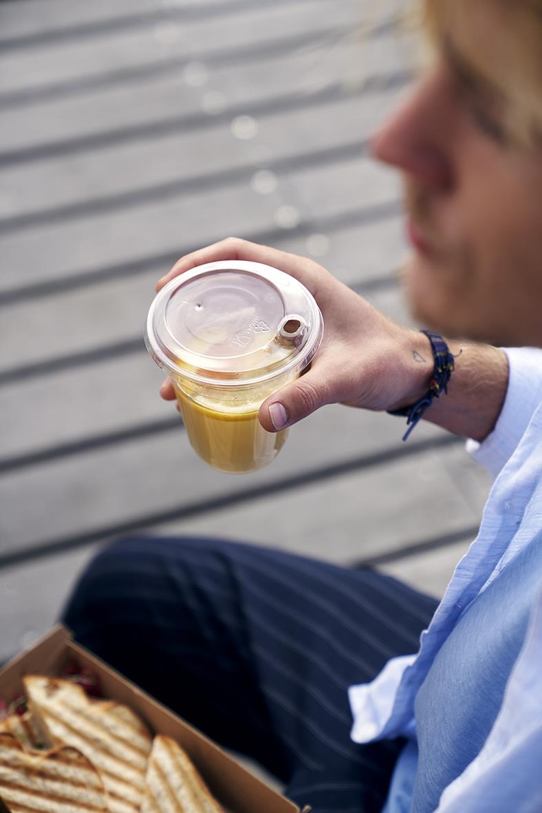 Person håller en genomskinlig pappersmugg med lock och pip, utomhus på en träbrygga