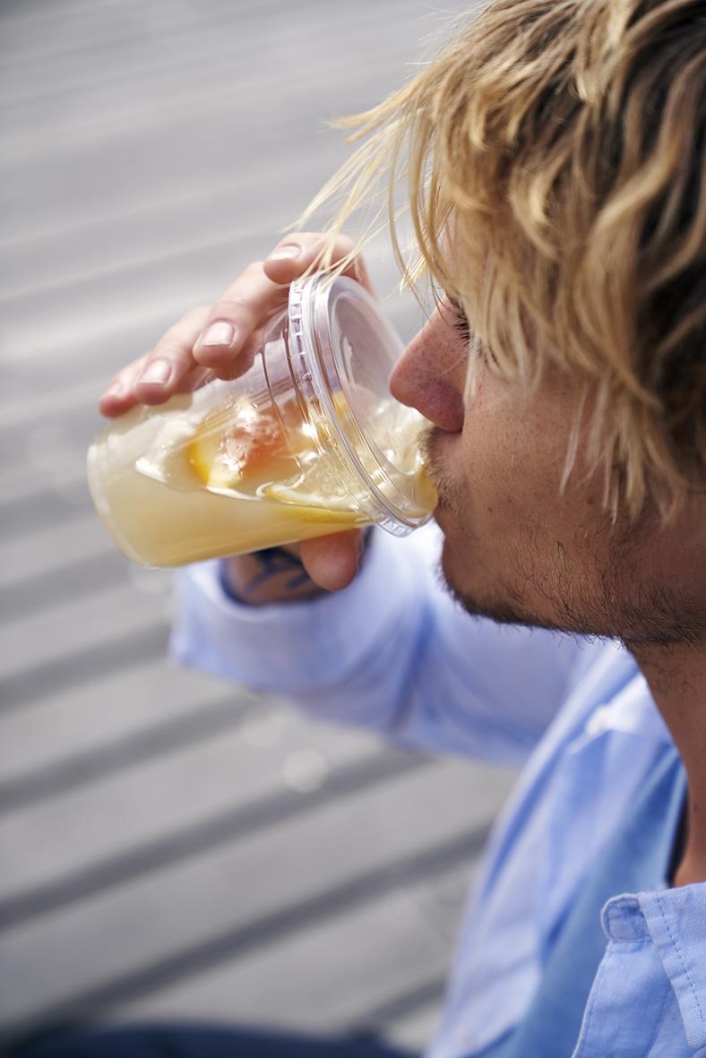 Man dricker från en klar pappersmugg med gul dryck, utomhus på en träbänk