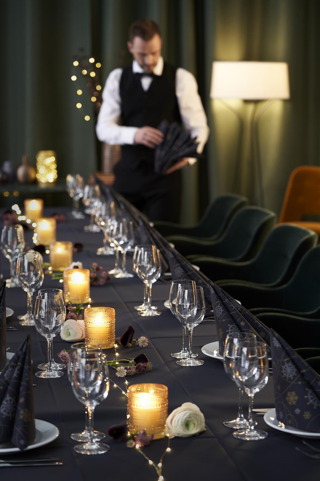 Elegant middagsduk i svart med glas, ljuslyktor och blommor på ett festbord, serveringspersonal i bakgrunden