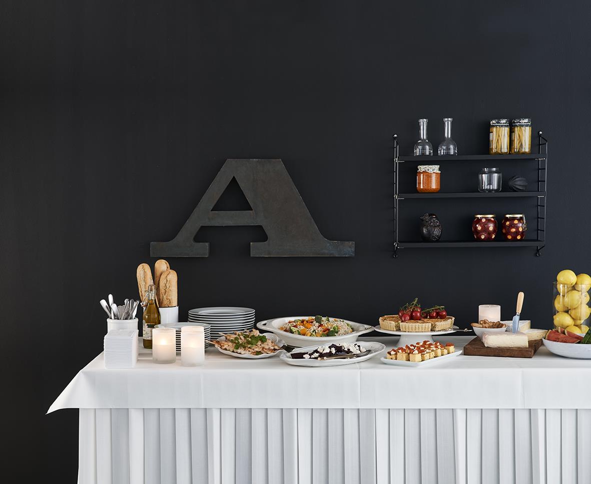 Bord med duk och mat, mörk vägg, dekorativa hyllor med burkar och ett stort bokstav A på väggen