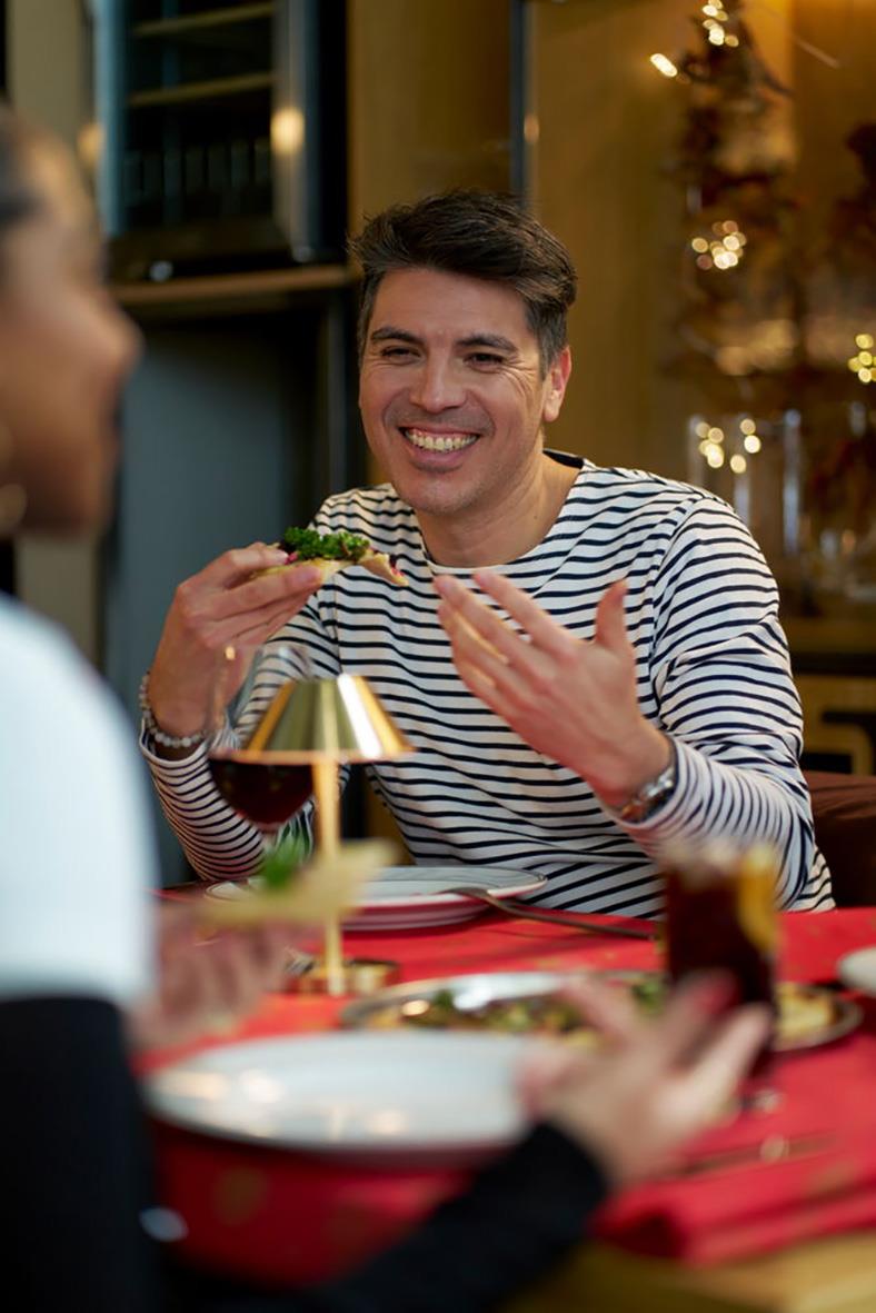 Glad man äter mat vid bord med röd duk, i ett mysigt inomhusmiljö med juldekorationer i bakgrunden