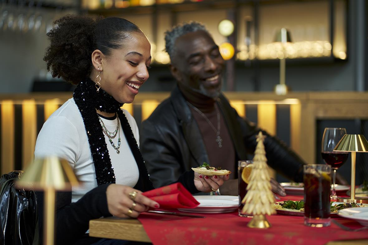 Två personer som äter och skrattar vid ett julbord med dekorationer och vin i en mysig inomhusmiljö