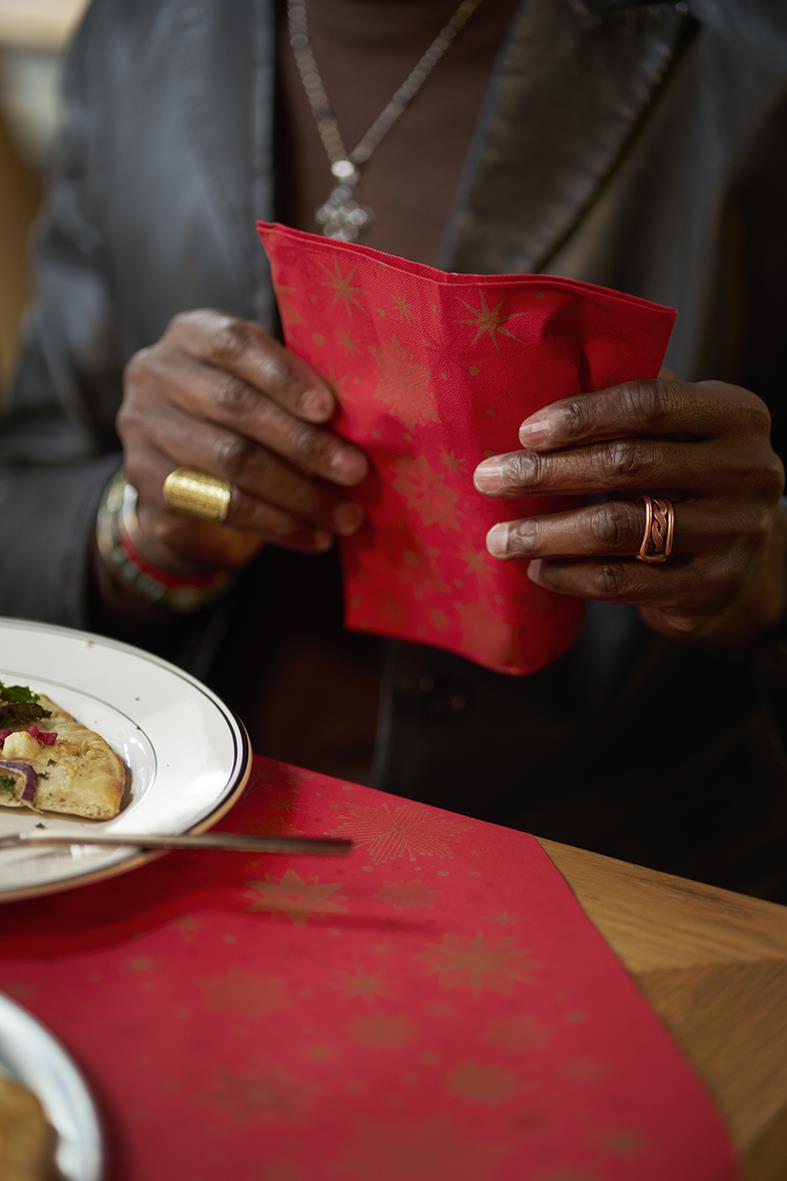 Person håller i en röd Dunisilk Stargazing Duni-duk med guldstjärnor, 84x84cm, vid ett bord med mat.