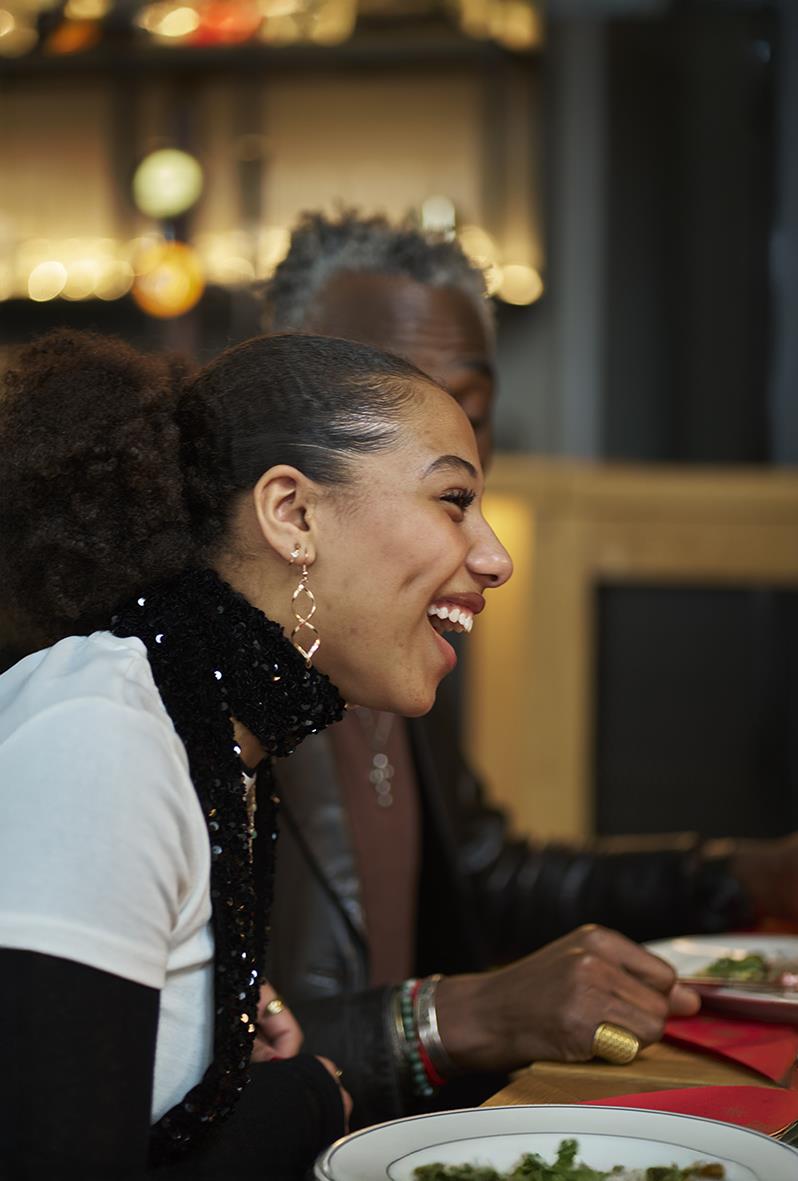 Två kvinnor skrattar och pratar vid ett bord i ett inomhusmiljö, en av dem har på sig en svart scarf och stora örhängen.