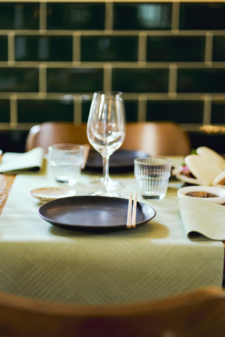 Bordduk med tallrik, glas, bestick och servetter i en restaurangmiljö med mörk vägg i bakgrunden