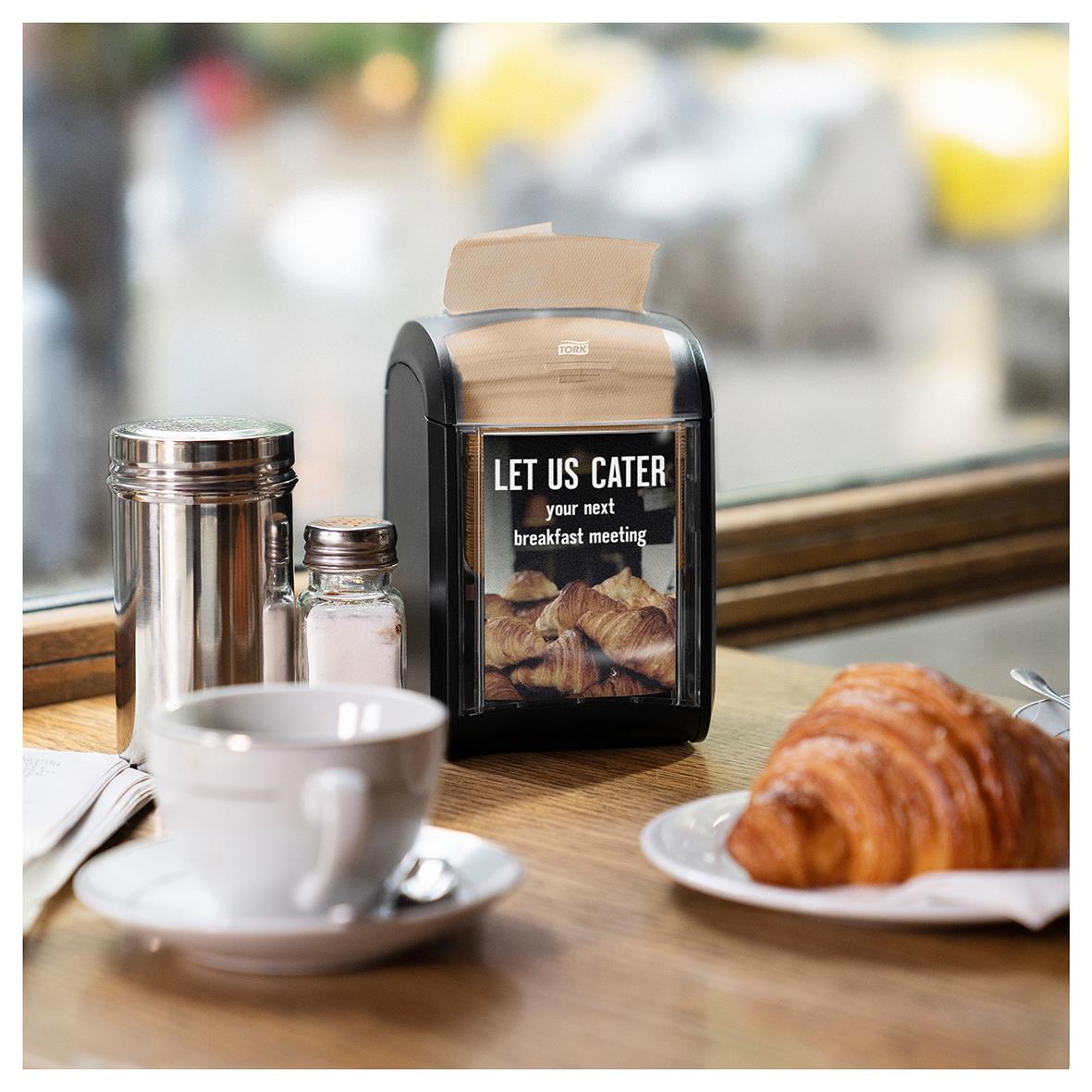 Dispenser för servetter Tork N14 2-lag naturfärgad, 16,5x21,3cm, placerad på ett bord med kaffe och croissant