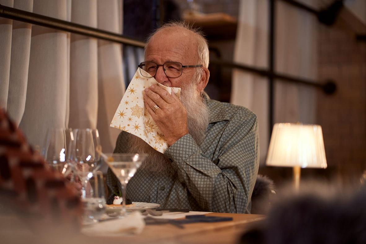 Äldre man med glasögon som nyser i en servett vid matbordet i ett mysigt rum