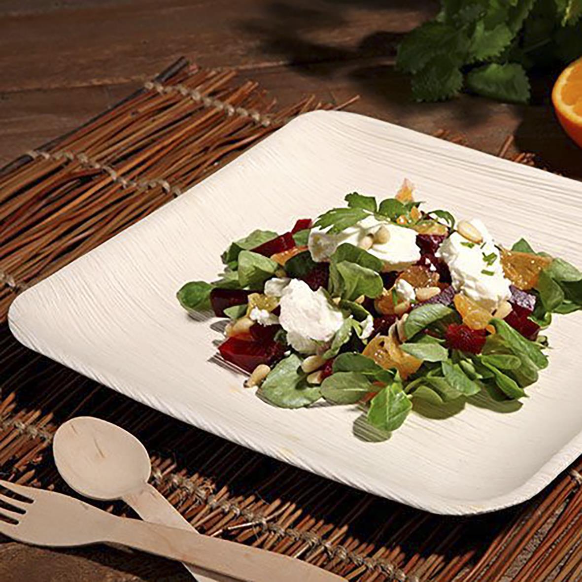 Engångstallrik av palmblad med kantig form och storlek 25,5x25,5 cm på träbord med bestick och sallad