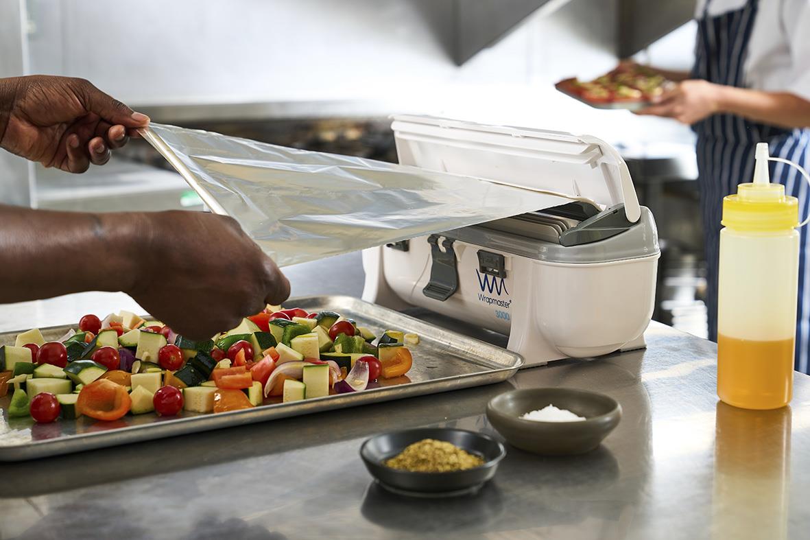Person använder Dispenser Wrapmaster 3000 för att linda in mat på en arbetsbänk med grönsaker och andra köksredskap