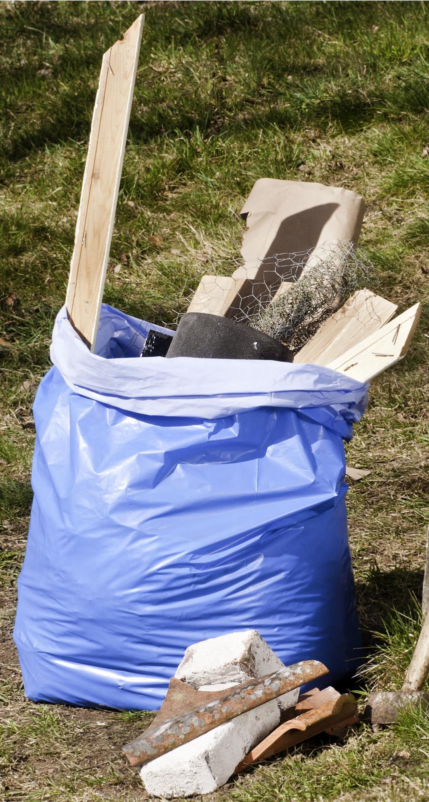 Blå sopsäck fylld med byggavfall, inklusive trä, metall och andra material, utomhus på gräs.