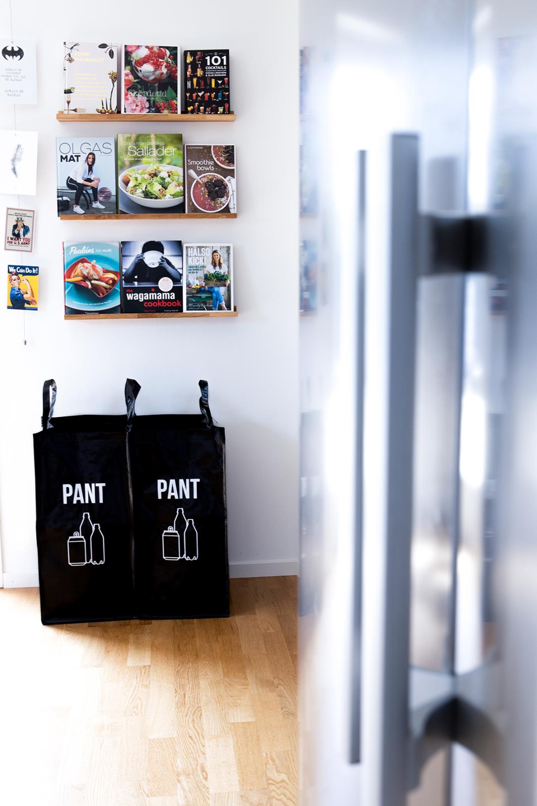 Två svarta pantpåsar med texten PANT och bild av en flaska, placerade mot en vit vägg med böcker ovanför.