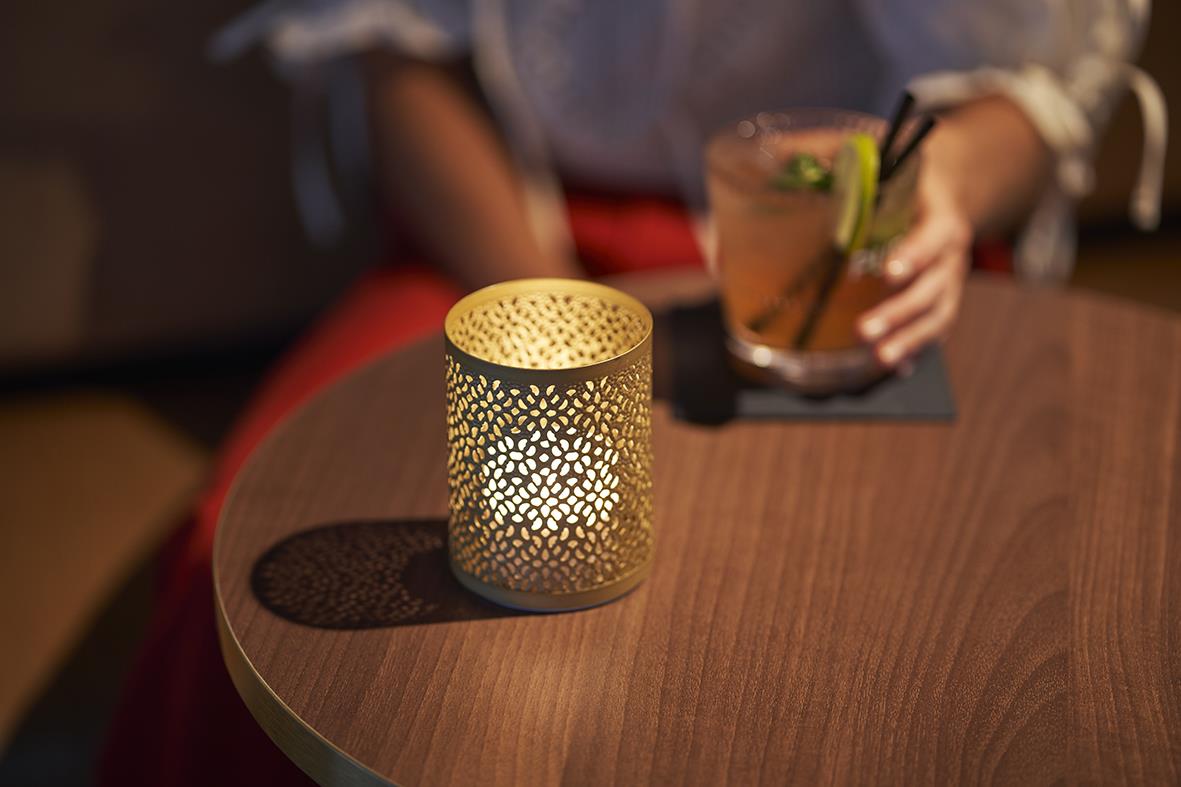 Guldig ljushållare Duni Bliss på ett runt bord med en person i bakgrunden som håller en drink