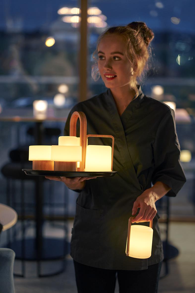 Kvinna bär en bricka med LED-ljushållare i bambu på kvällen, utsikt över vatten och båtar i bakgrunden