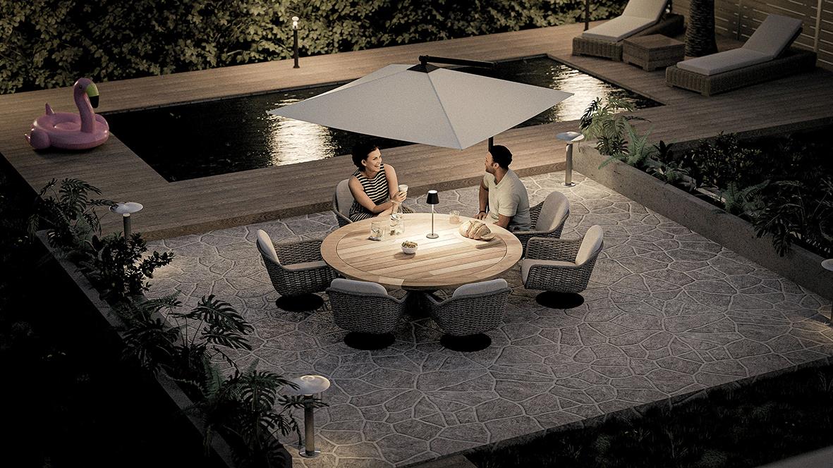 " personer sitter runt ett runt bord med LED-lampa utomhus på kvällstid, omgiven av träd och utemöbler"