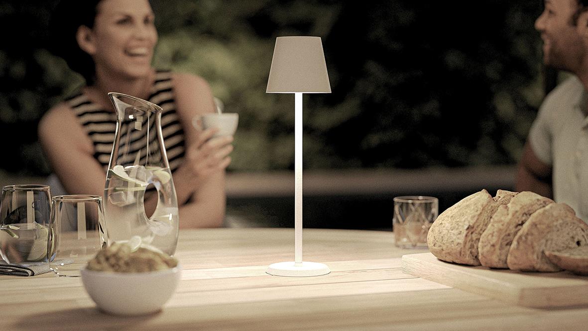 Bordslampa LED Securit Feline Vit på ett bord med bröd och drycker, utomhus med glada personer i bakgrunden