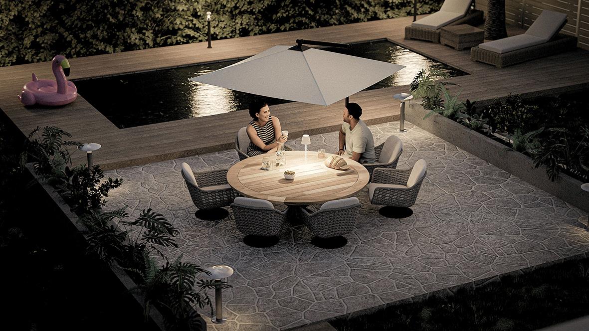 Två personer sitter vid ett runt bord med en vit parasoll på en uteplats med pool och träd i bakgrunden