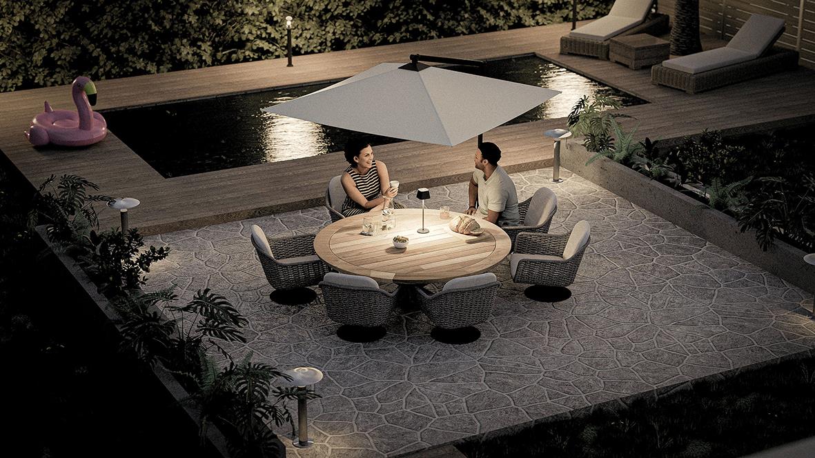 Två personer sitter vid ett runt bord med en stor vit parasoll, utomhus på kvällstid, omgiven av träd och utemöbler