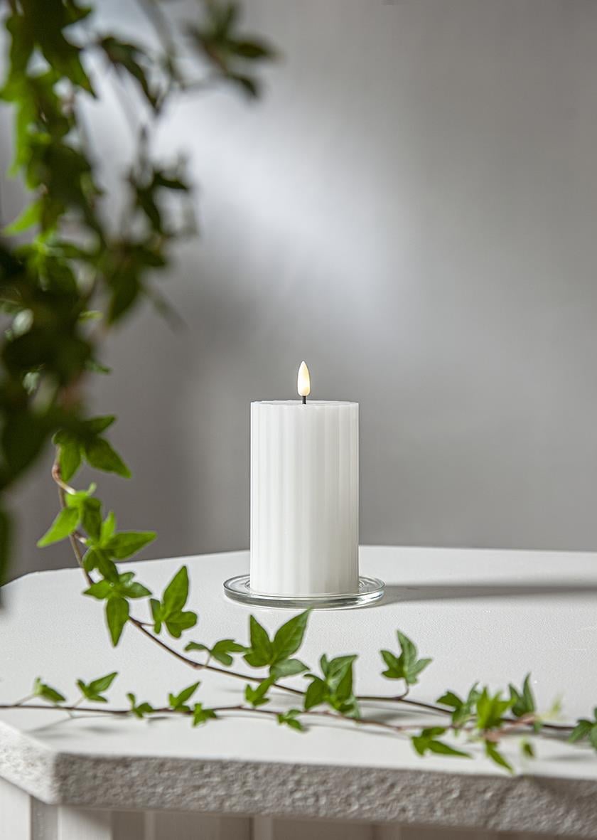 Vitt LED blockljus med flammemönster på en vit yta med gröna växter i förgrunden