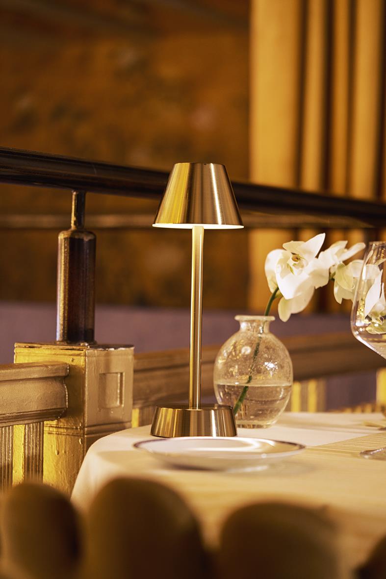 Bordslampa LED Duni Zelda i mässing placerad på ett bord med vita blommor och glas i bakgrunden