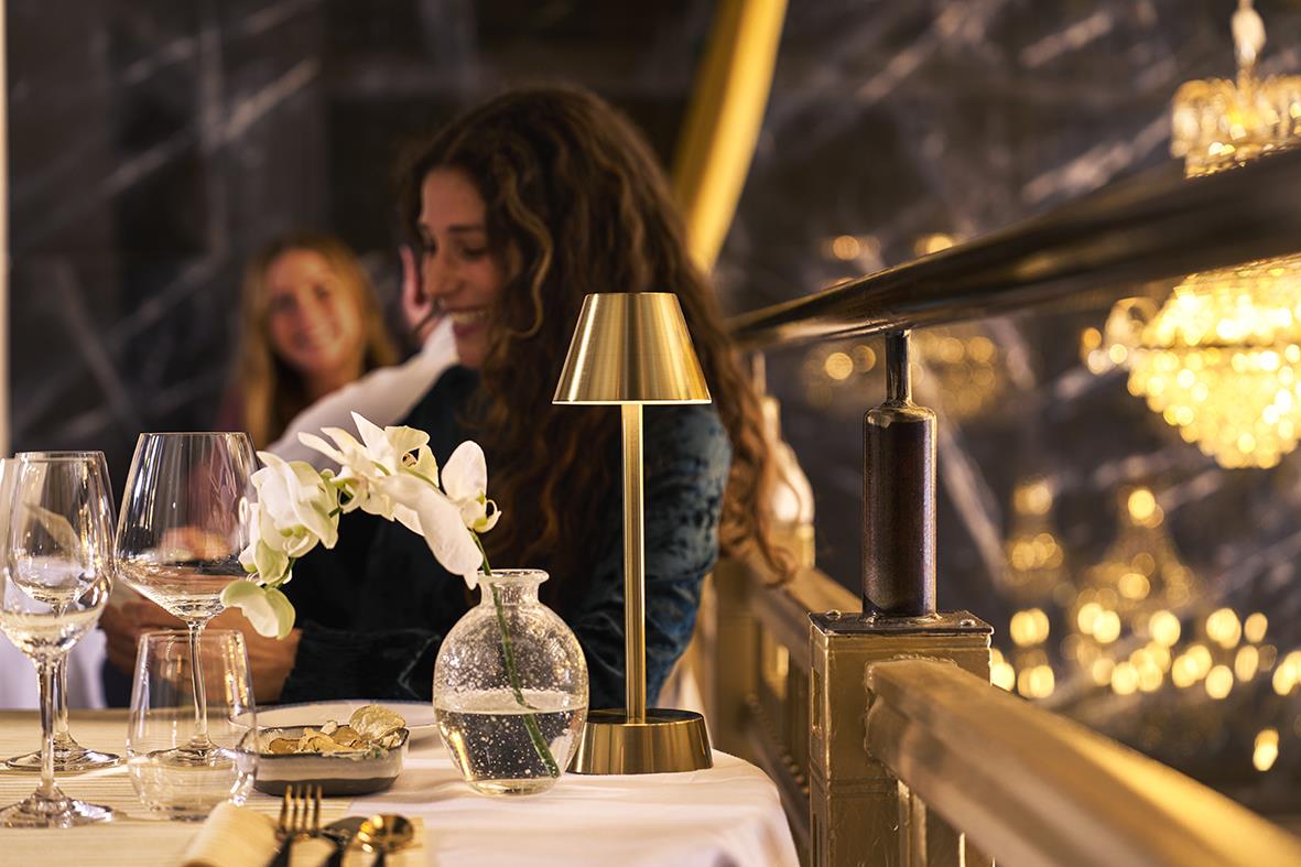Bordslampa LED Duni Zelda Mässing på ett bord med glas och blommor i en elegant restaurangmiljö