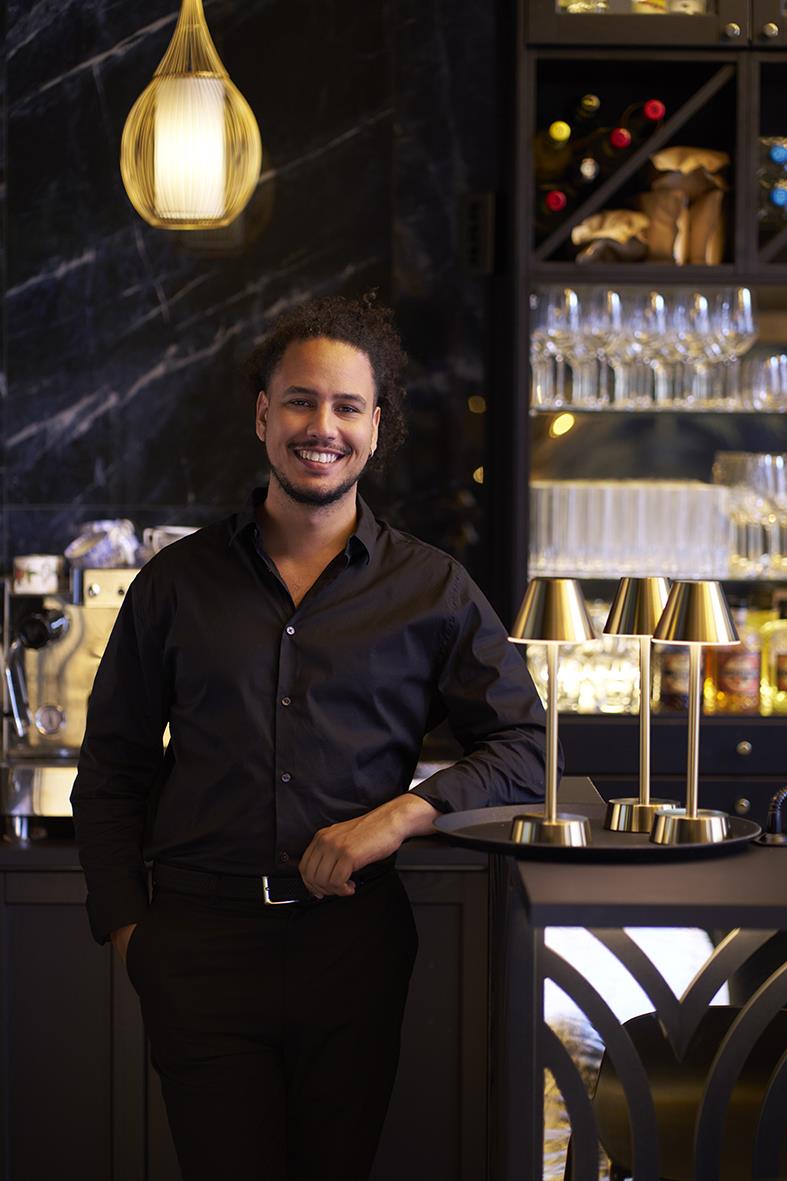 Man i svart skjorta står vid baren med guldlampor och bordslampa LED Duni Zelda Mässing i bakgrunden