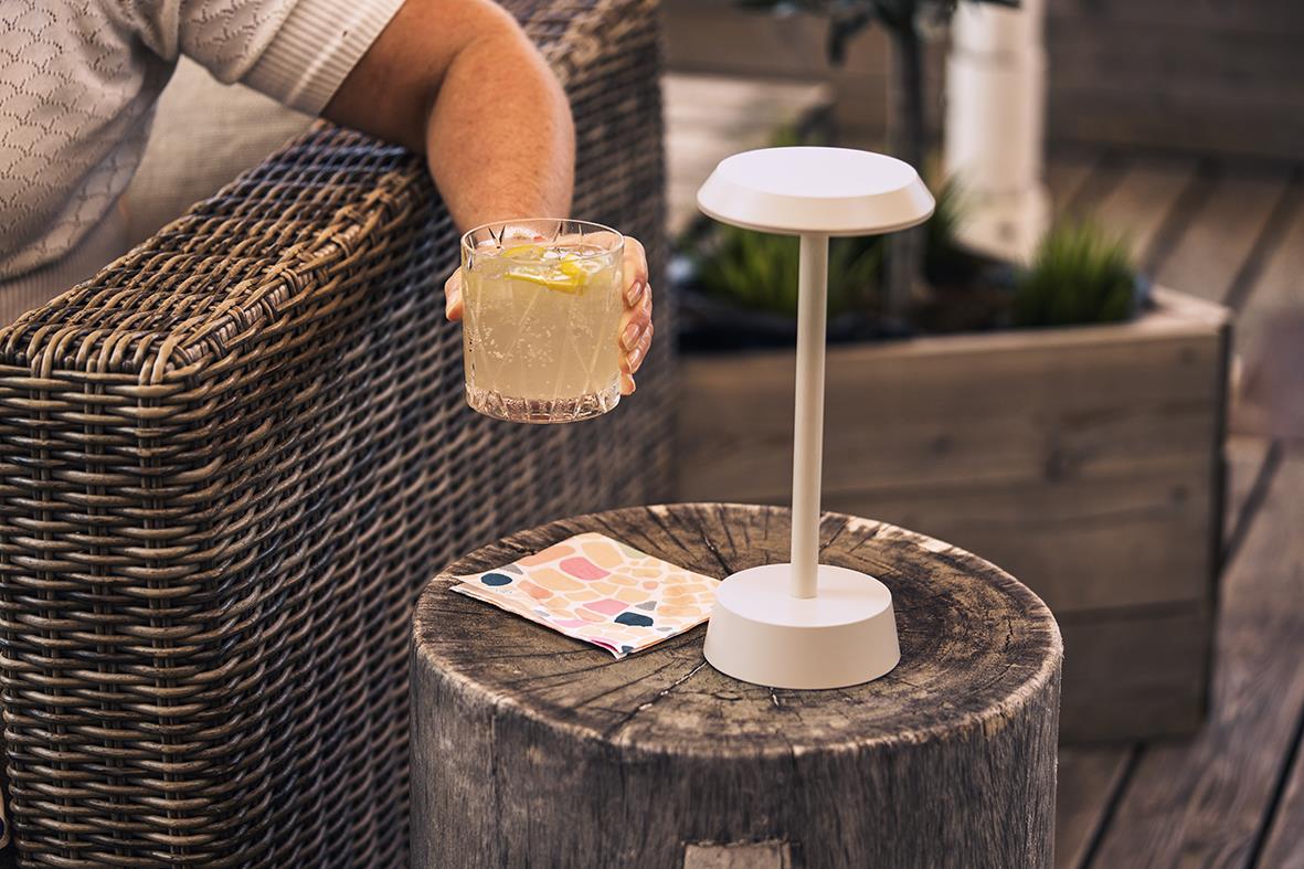 Bordslampa LED Duni Nour Sand på ett träbord bredvid en person som håller en drink