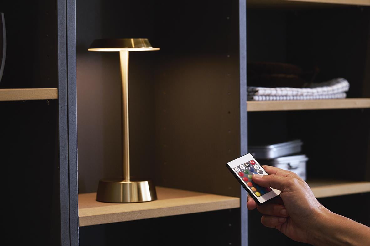 Bordslampa LED Duni Nour i mässing på hylla, person justerar ljusstyrka med fjärrkontroll