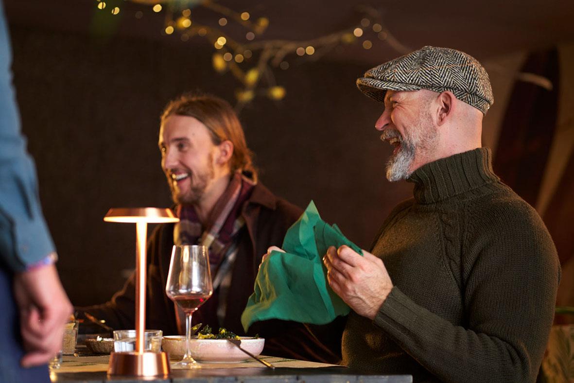 Två män som pratar och skrattar vid ett bord med en LED-lampa Duni Nour Rose Gold i bakgrunden