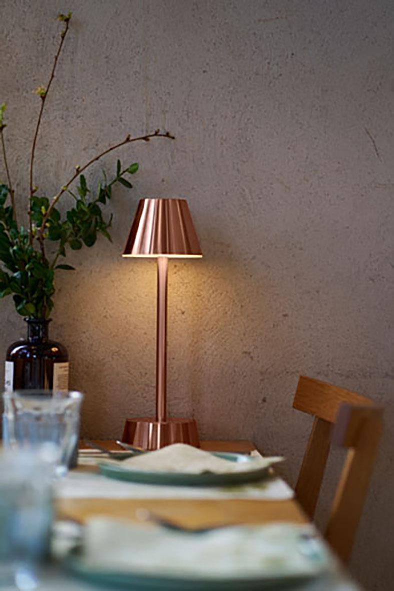 Bordslampa LED Duni Zelda Rose Gold på ett bord med duk och glas, nära växt och vägg