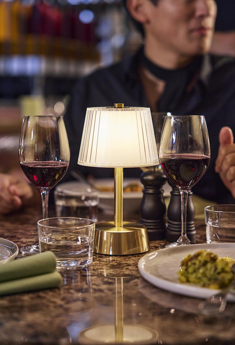 Bordslampa LED Duni Jay Guld på ett bord med glas vin och mat, i bakgrunden en person i mörk klädsel