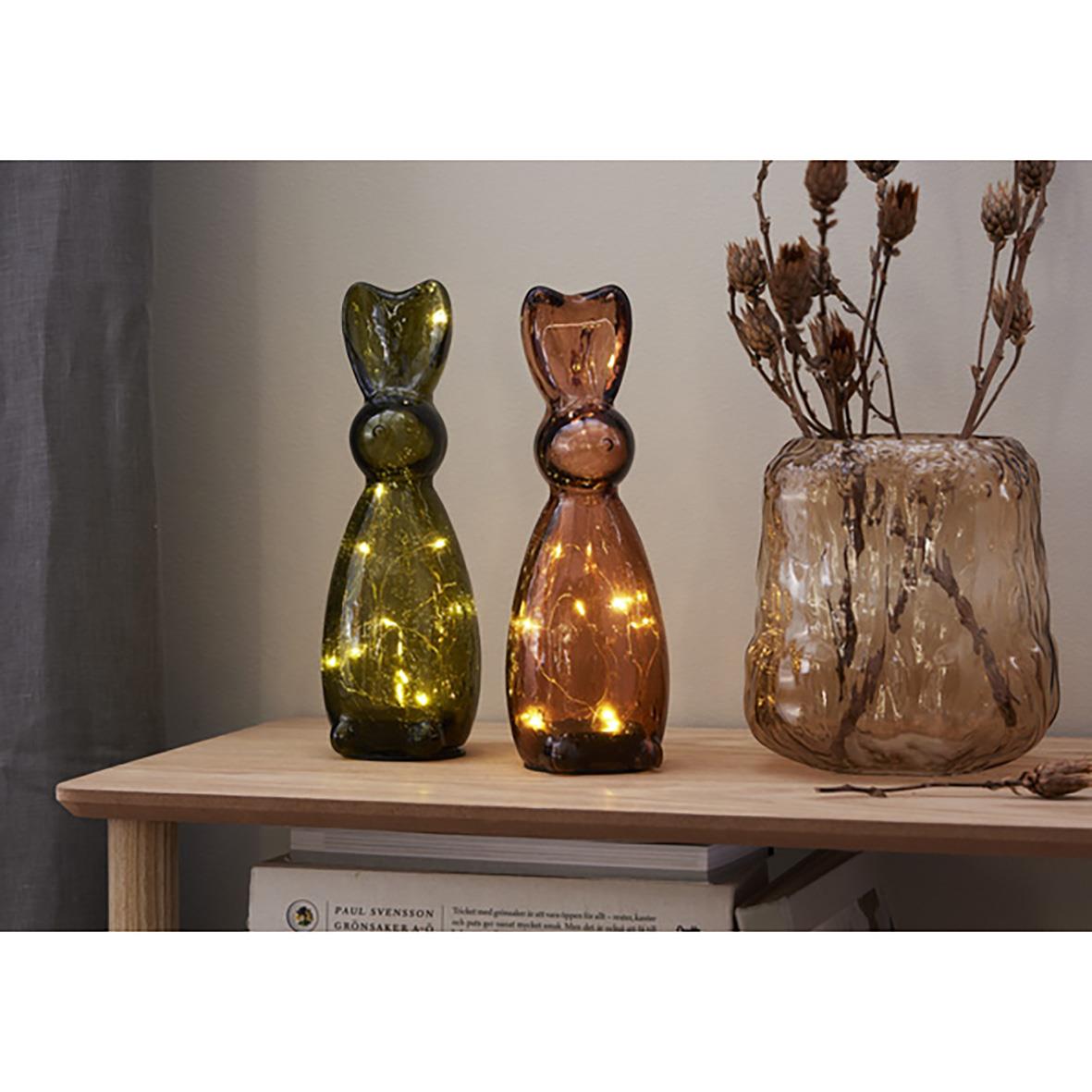 Två glasdekorationer i form av katter, en grön och en rosa, samt en stor glasvas med torkade växter på ett träbord