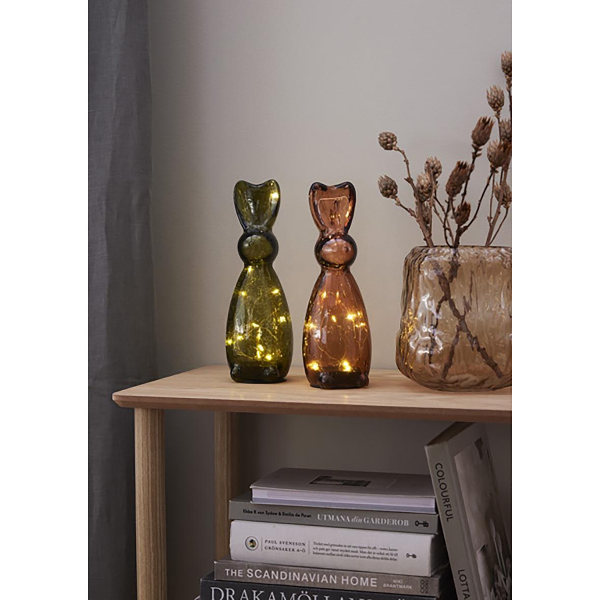 Två glasdekorationer i form av kattöron, en i grönt och en i brunt, placerade på ett träbord bredvid en vas med torkade växter