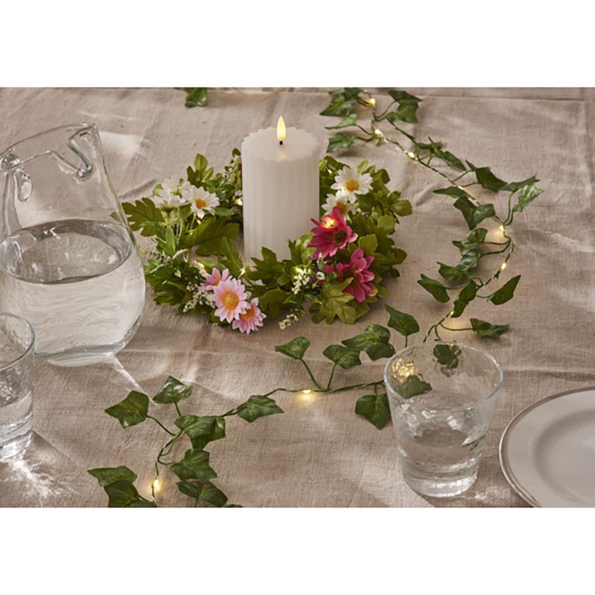 Ljusslinga Ivy runt ett vitt ljus med blommor och grönt runt ett bord